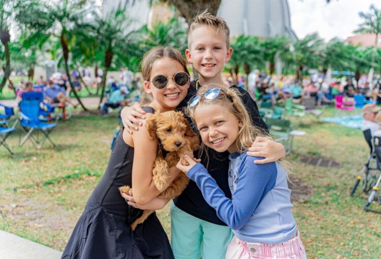 Three young siblings smile at the camera with their puppy being held in the center