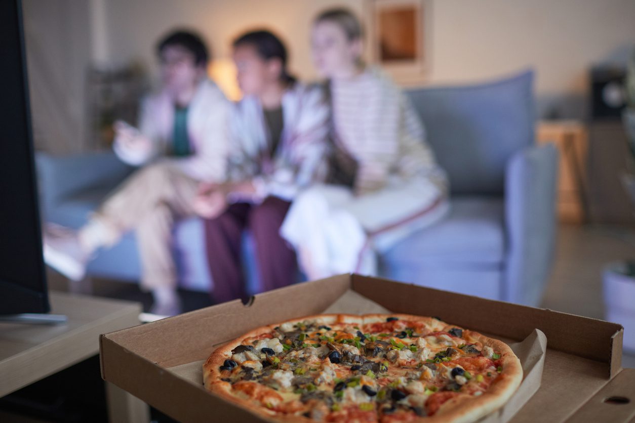 Blurred group of friends watching TV at home lit by blue light, focus on pizza in foreground, copy space